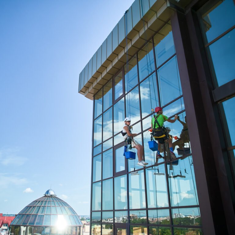 Industrial mountaineering. Male washers hanging on ropes and using cleaning tools while washing building windows. Two men window cleaners in safety helmets working together at high-rise building.