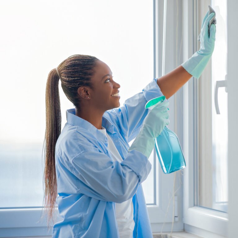 Professional housekeeping, cleaning service. Profile portrait of young african american woman housekeeper cleaning windows at apartment, using spray and cloth, wearing rubber gloves