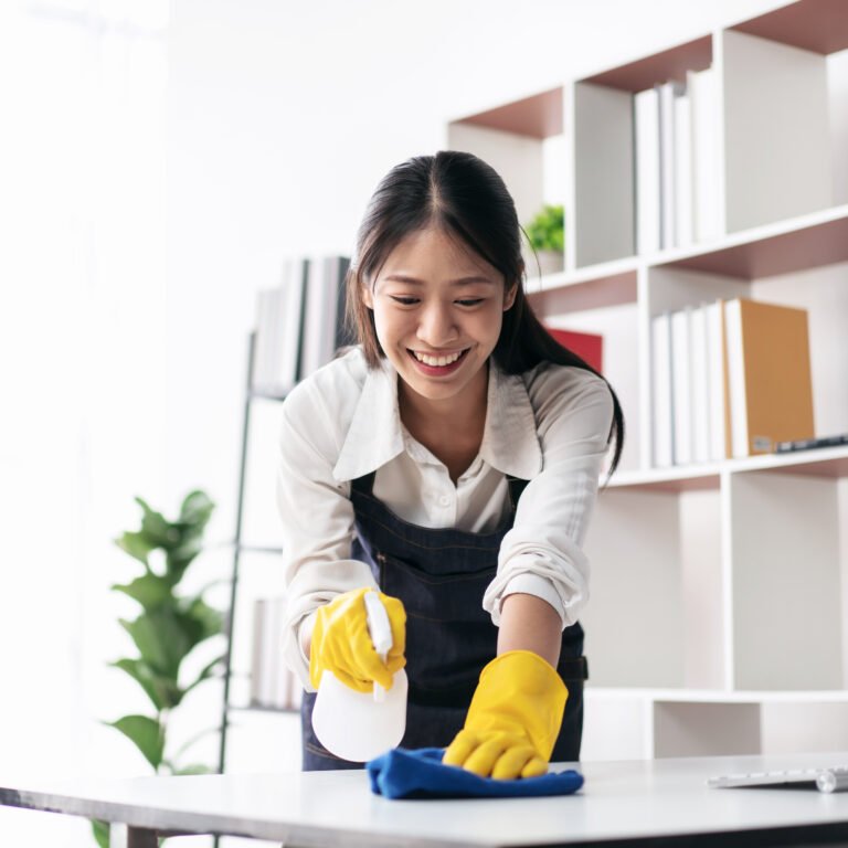 Housewife in apron wearing gloves to spraying hygiene spray on computer and using microfiber fabric to wiping cleanup the table while working and cleaning furniture in the house.